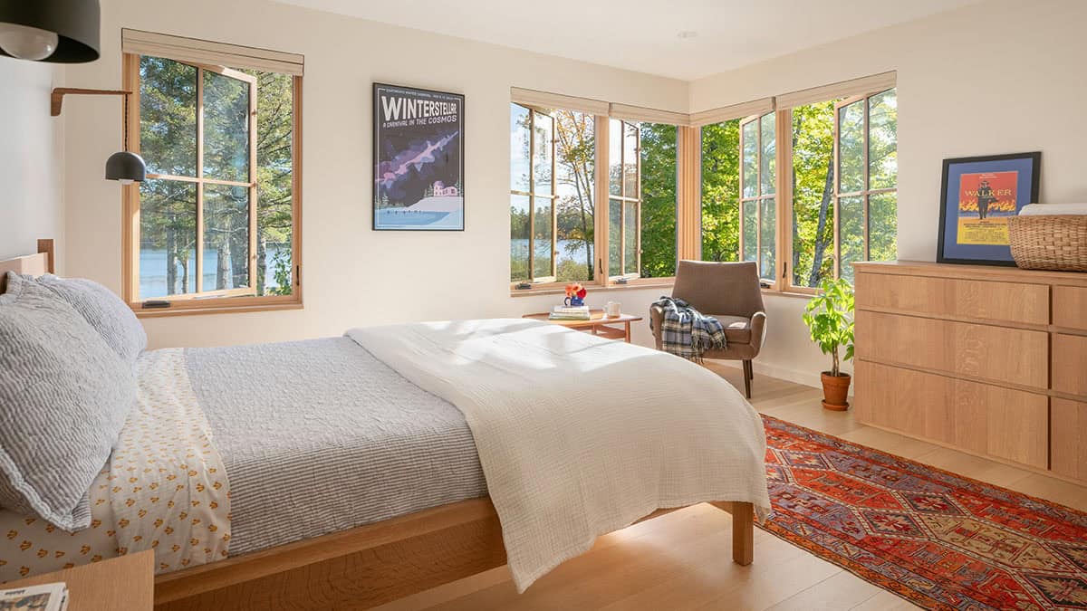 Bright bedroom with a wood bed frame, patterned rug, and corner windows overlooking trees
