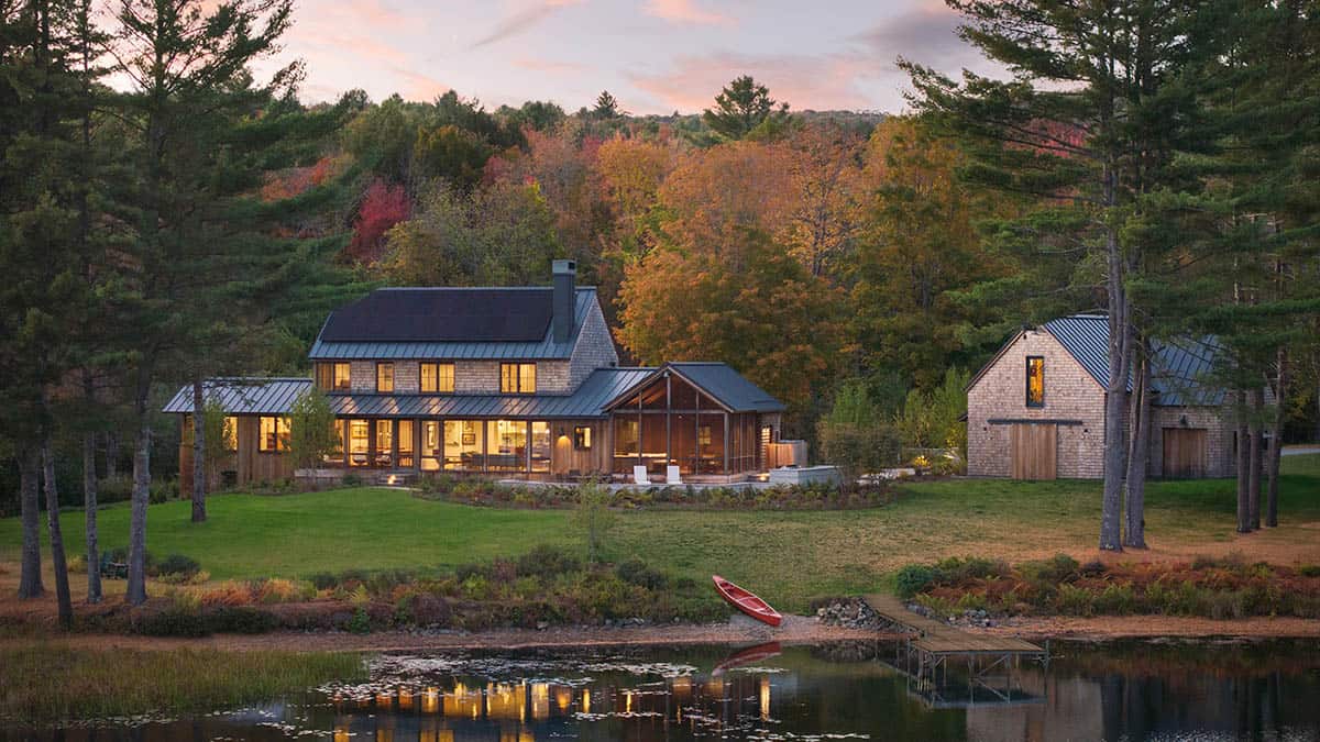 Twilight view of a shingled lake house and matching barn, glowing warmly beside a canoe on the shore