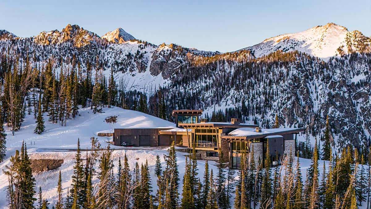 Modern mountain home nestled in snowy pine forest with rugged peaks behind, aerial view