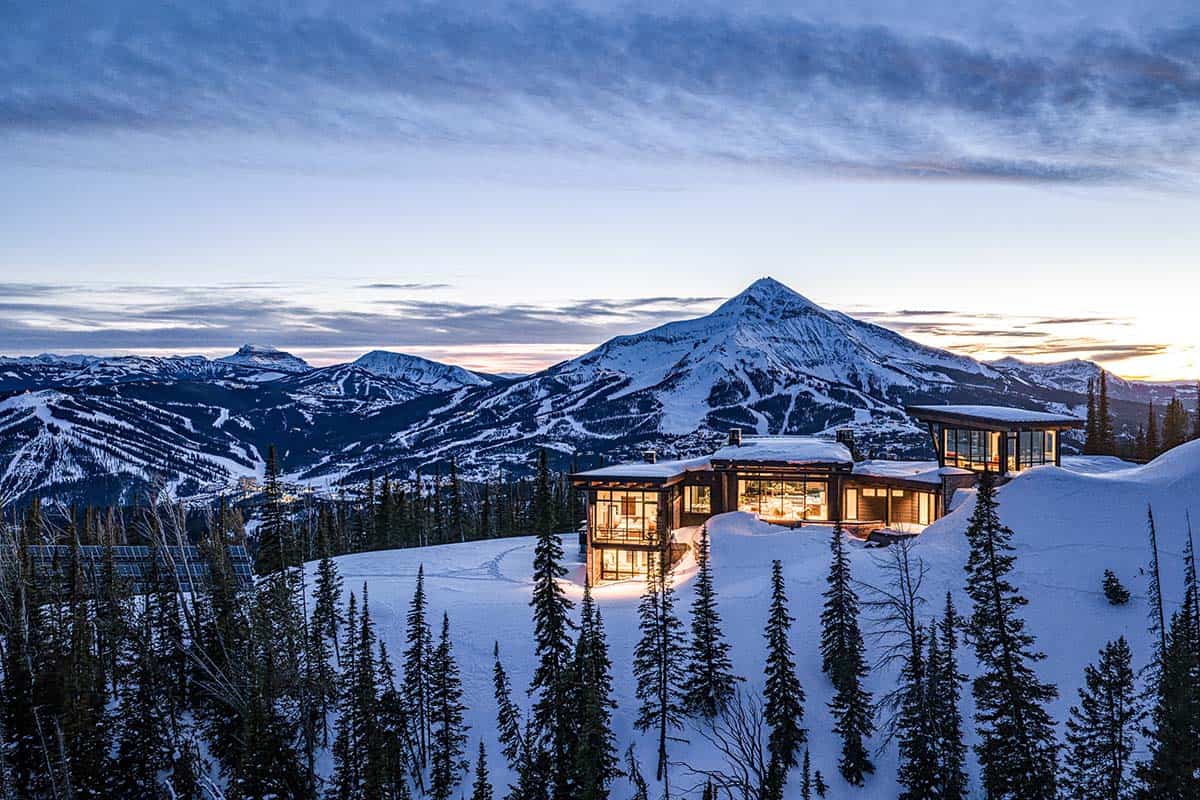 Illuminated mountain house at twilight with snow-capped peak and ski runs in the distance