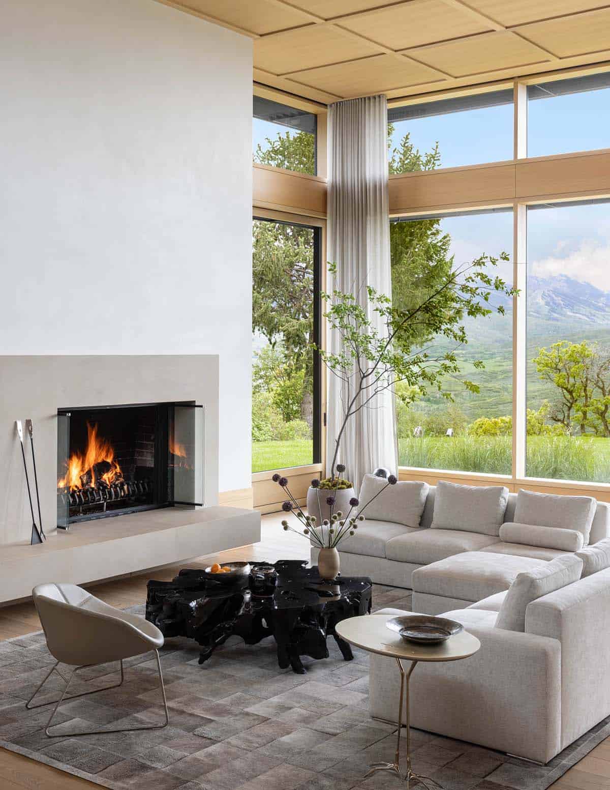 Living room with lit fireplace, gray sectional, and tall windows framing green valley