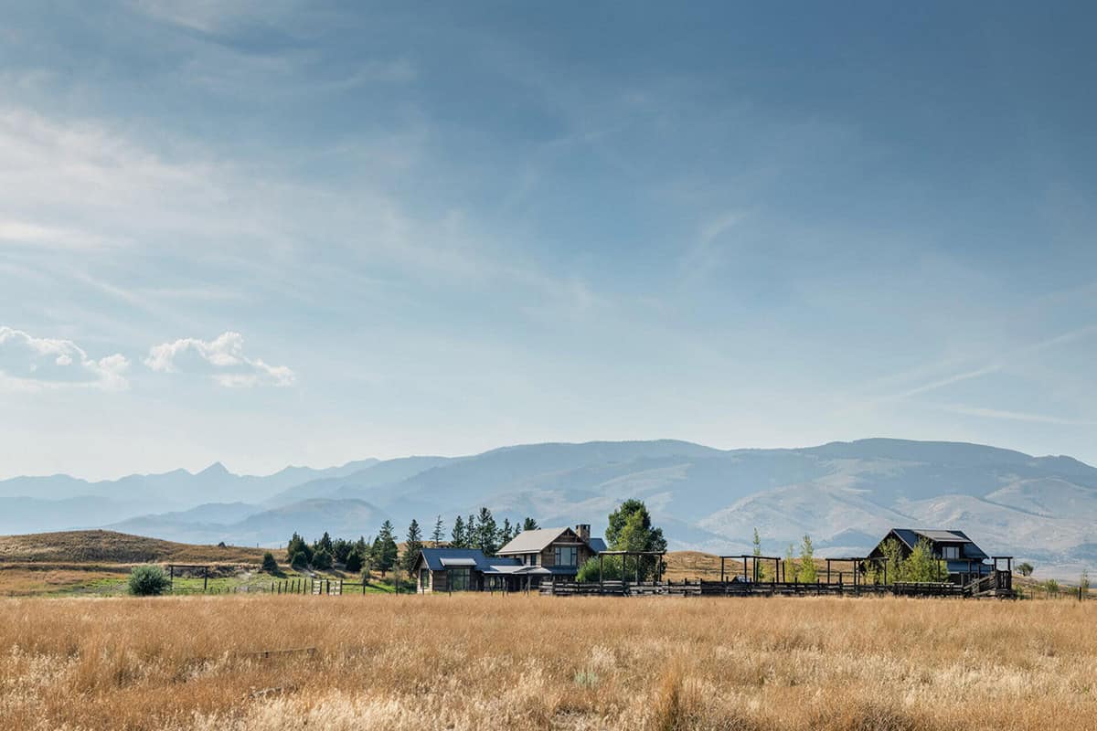 Wide shot of ranch buildings sitting low in a golden grass field beneath mountains