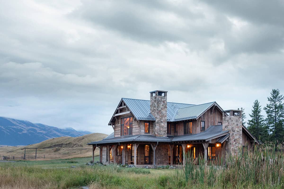 Two-story timber ranch house with stone chimneys at dusk, mountains in the distance