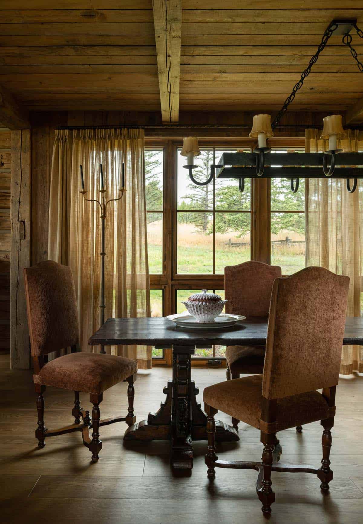 Rustic dining table with upholstered chairs beneath an iron chandelier by a large window