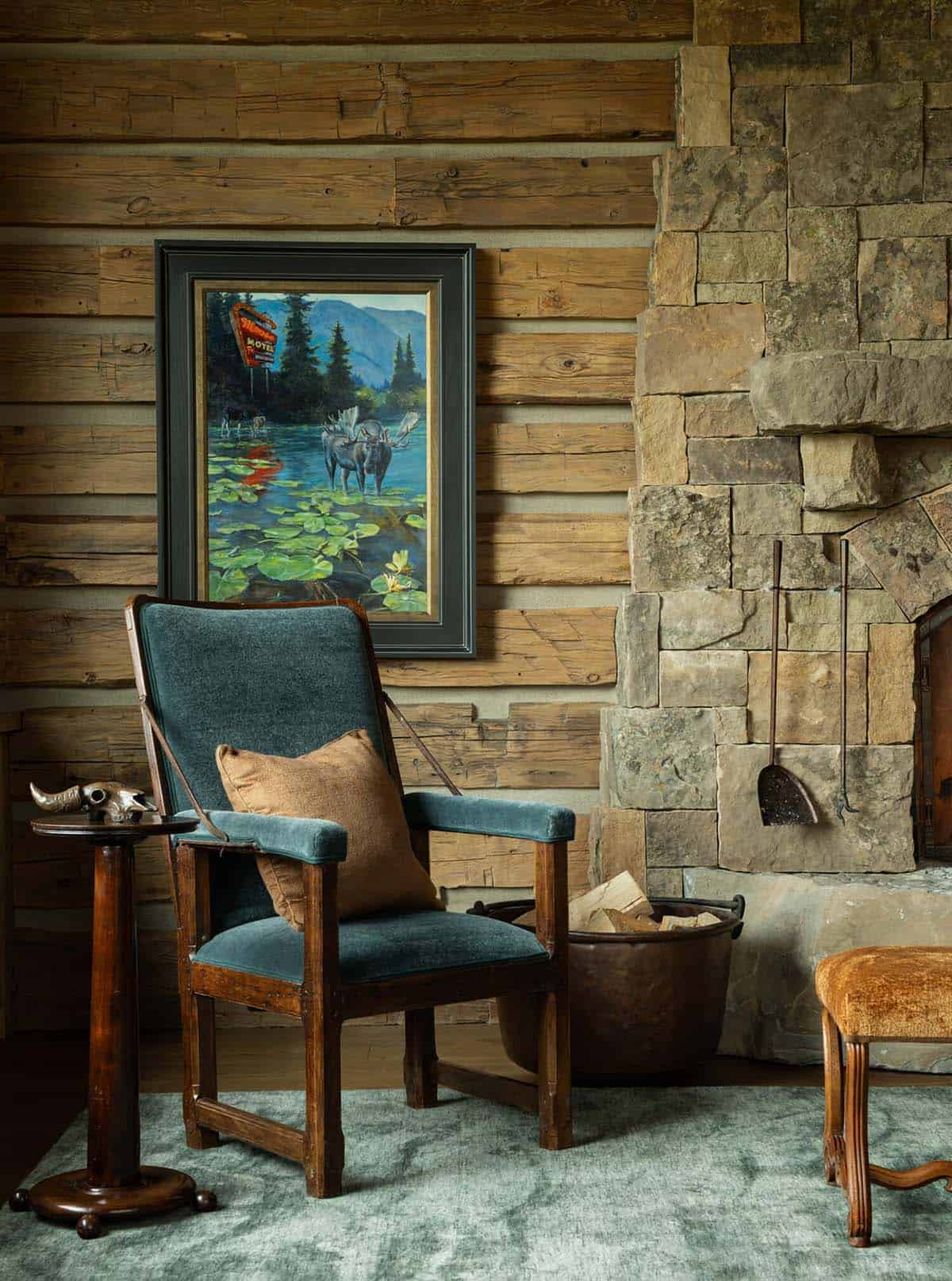 Teal armchair by a stone fireplace with a moose painting and log-filled copper basket