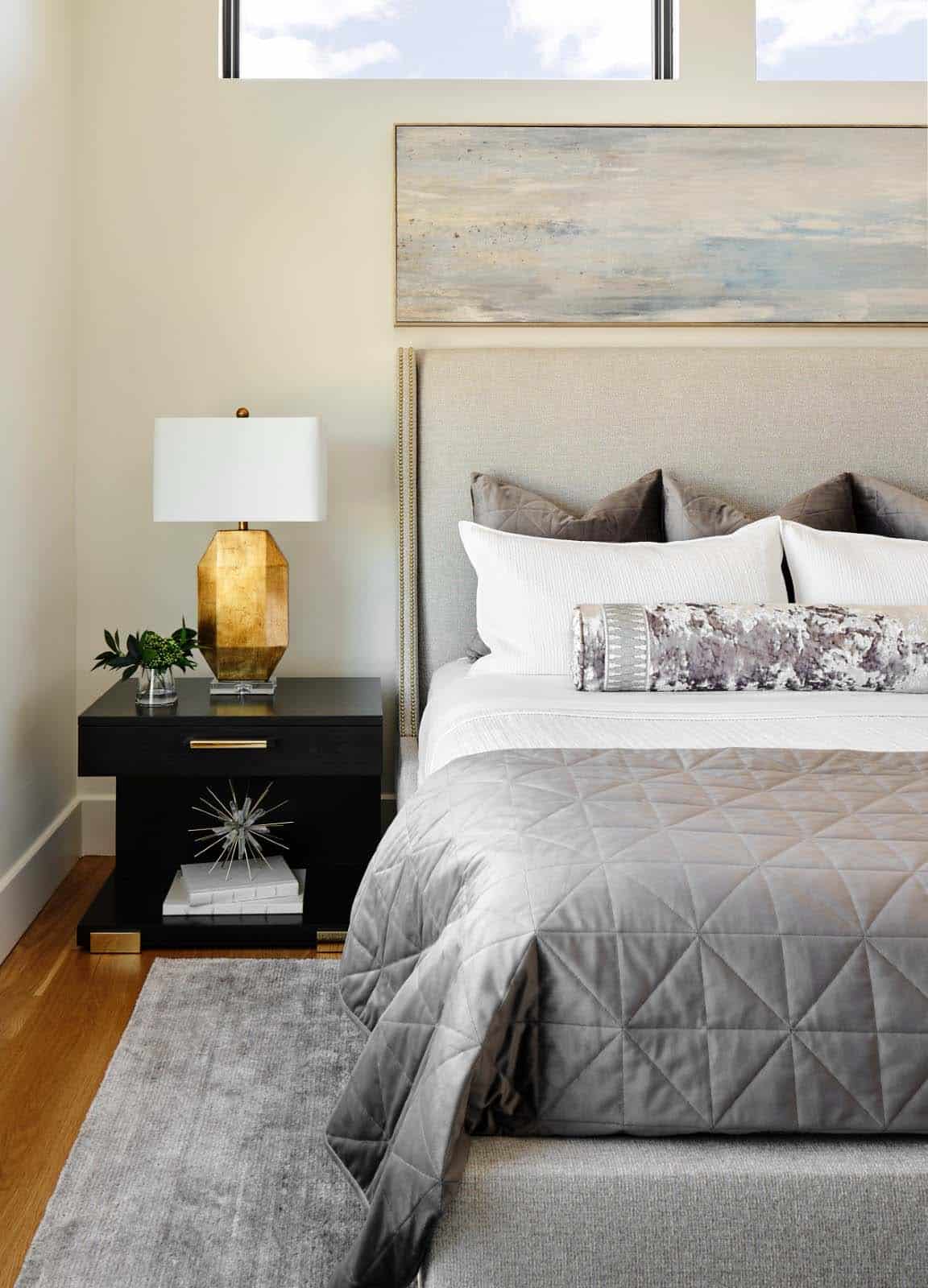 Bedside detail with brass geometric lamp, black nightstand, and quilted gray bedding
