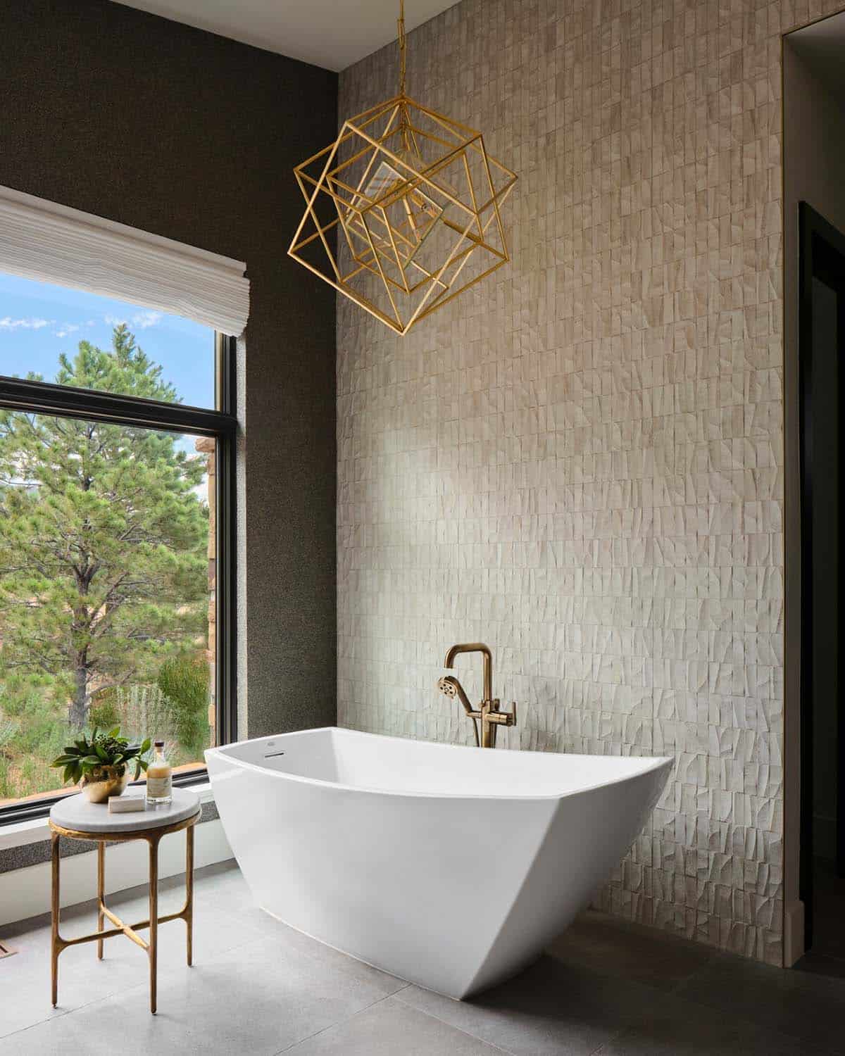 Freestanding white soaking tub beneath geometric brass pendant light and textured tile wall