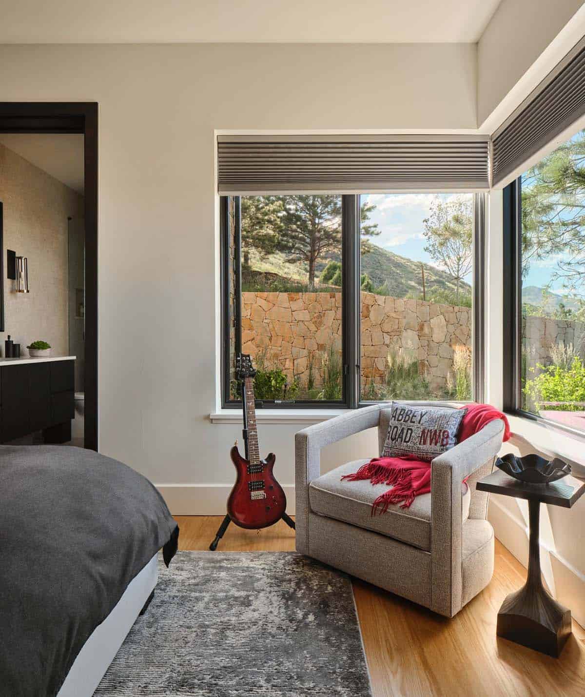 Corner reading nook with gray swivel chair, red guitar, and stone wall view through windows
