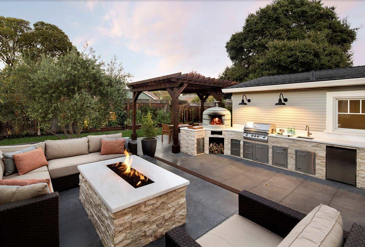 Napa-style backyard patio with a stacked stone fire pit and wicker sectional seating in the foreground, and a stone outdoor kitchen with a built-in grill and wood-fired pizza oven