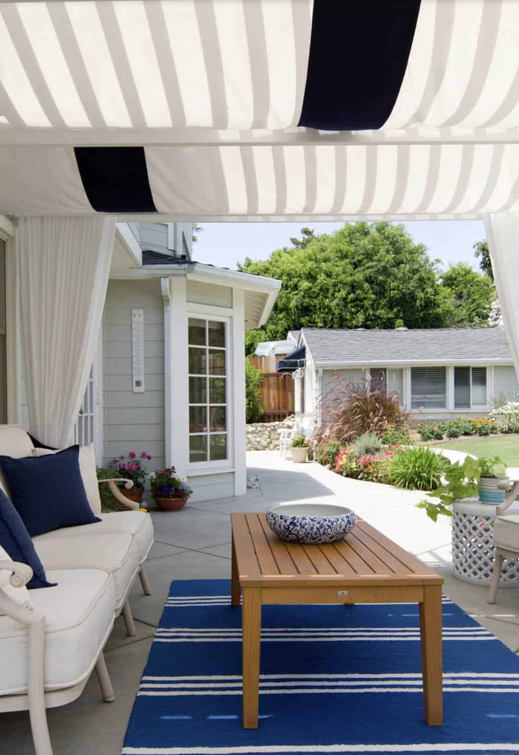 Covered patio lounge shaded by a navy and white striped fabric canopy, with a cream sofa, wood coffee table, and matching striped outdoor rug
