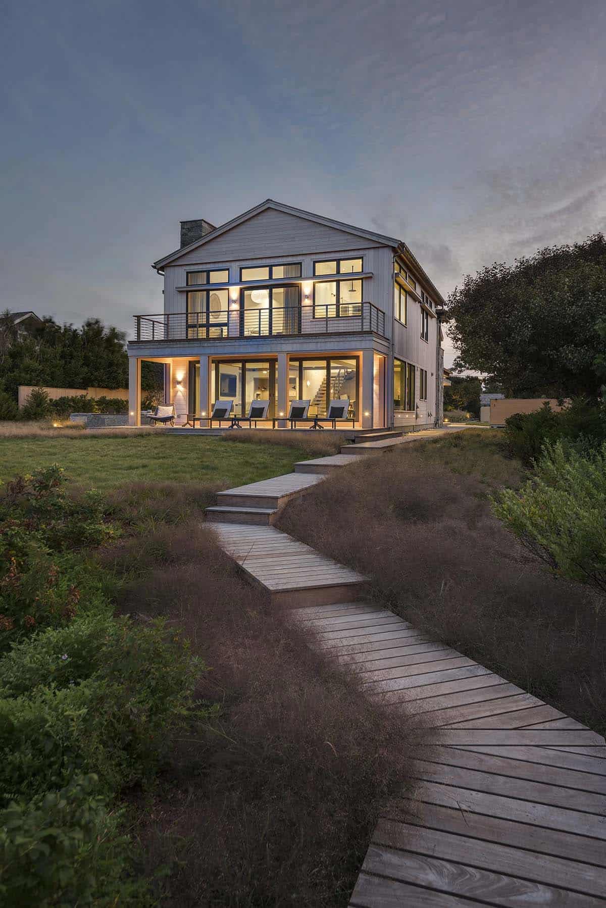 Two-story beach house with cedar shingle siding and gabled roof, lit up at dusk, viewed from a wooden boardwalk path through dune grass