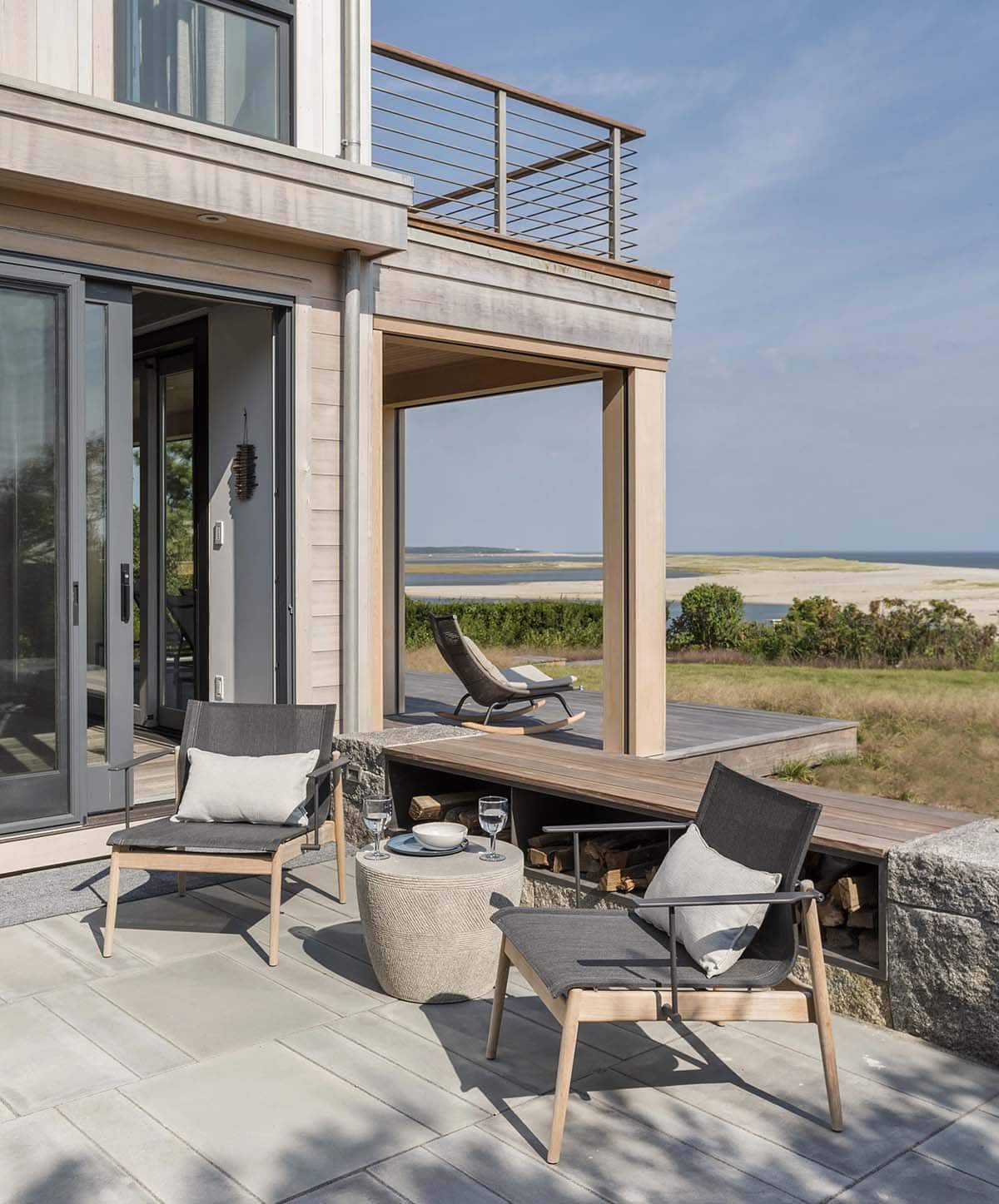 Covered patio with dark outdoor lounge chairs and a round stone side table, overlooking a wood-clad breezeway and ocean view
