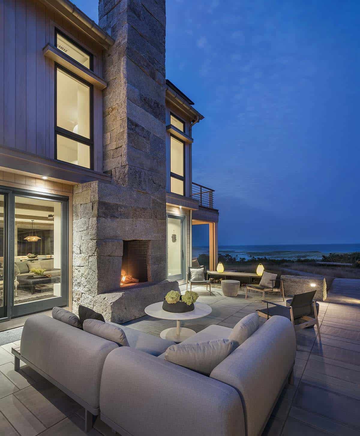 Outdoor stone fireplace with lit fire beside a gray sectional sofa and lounge chairs on a stone patio at dusk, ocean in background