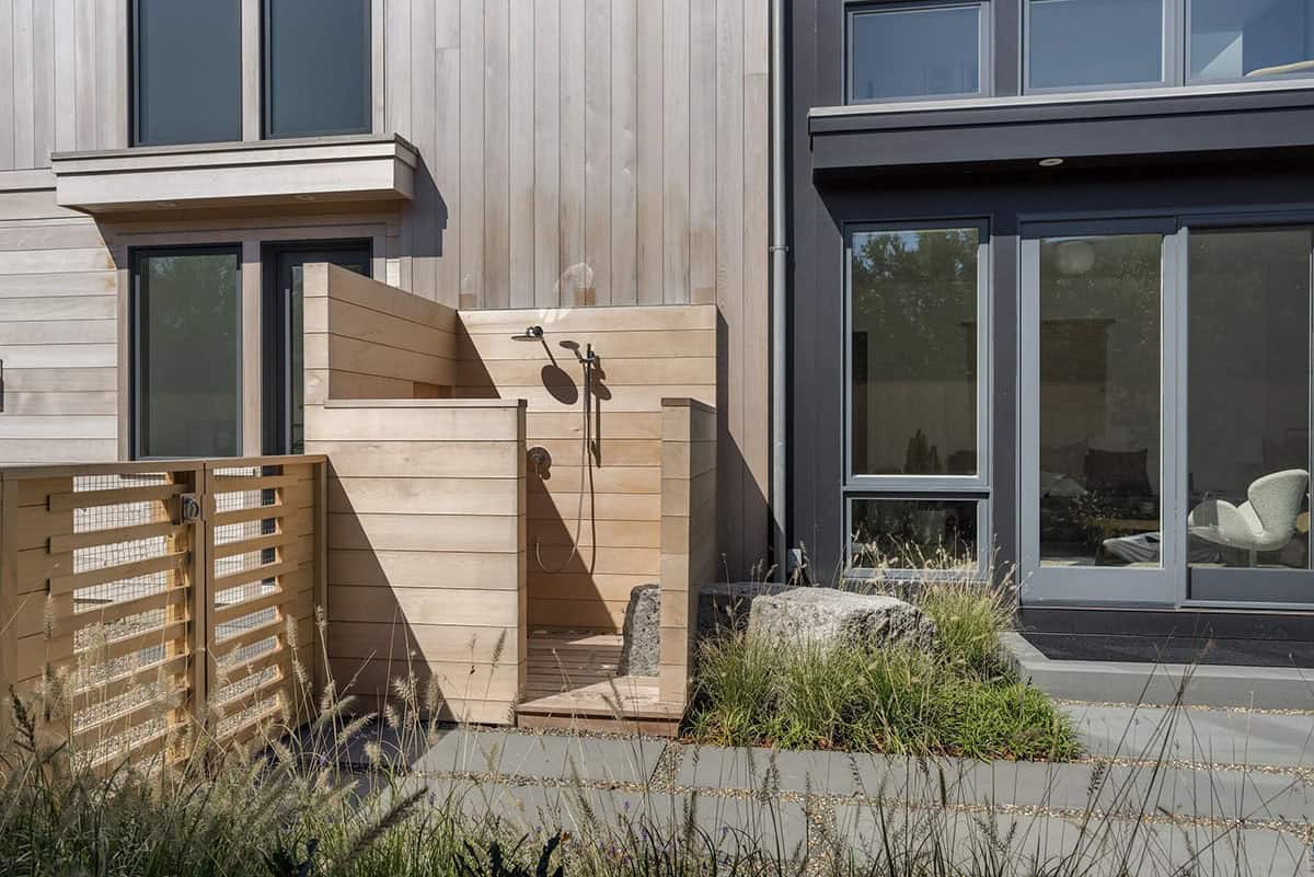 Outdoor wood-paneled shower enclosure beside the home's cedar and dark-framed exterior, with boulders and ornamental grasses
