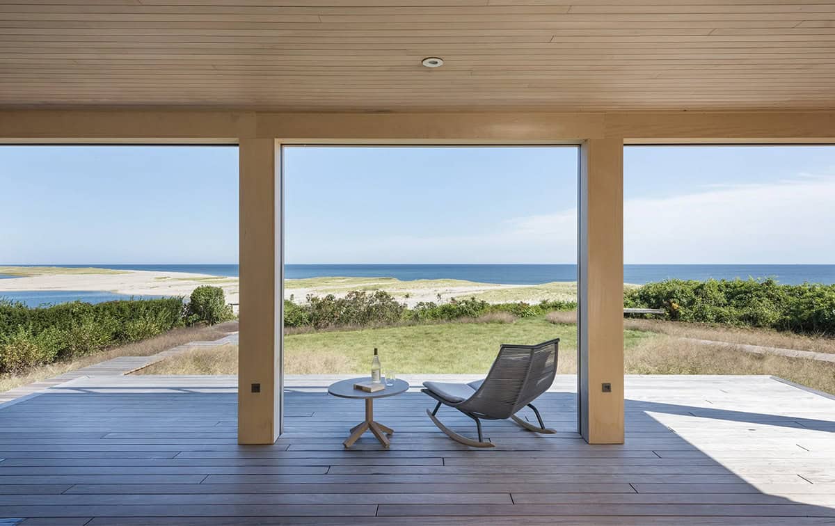 Covered wood deck framed by three large open bays with a rocking chair and small table, overlooking dunes and the ocean
