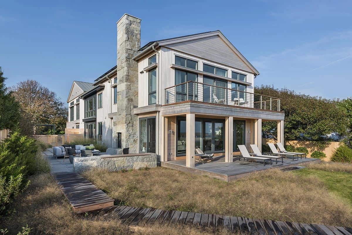 Two-story shingled beach house with stone chimney, cable-rail balcony, and covered lower deck with lounge chairs, seen from the lawn