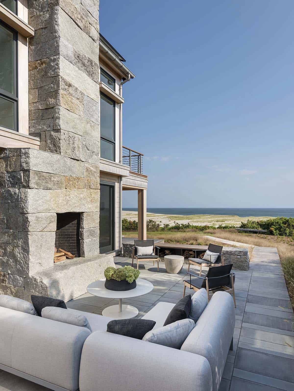 Stone chimney and covered patio with gray outdoor sofa, chairs, and built-in firewood storage, overlooking the ocean