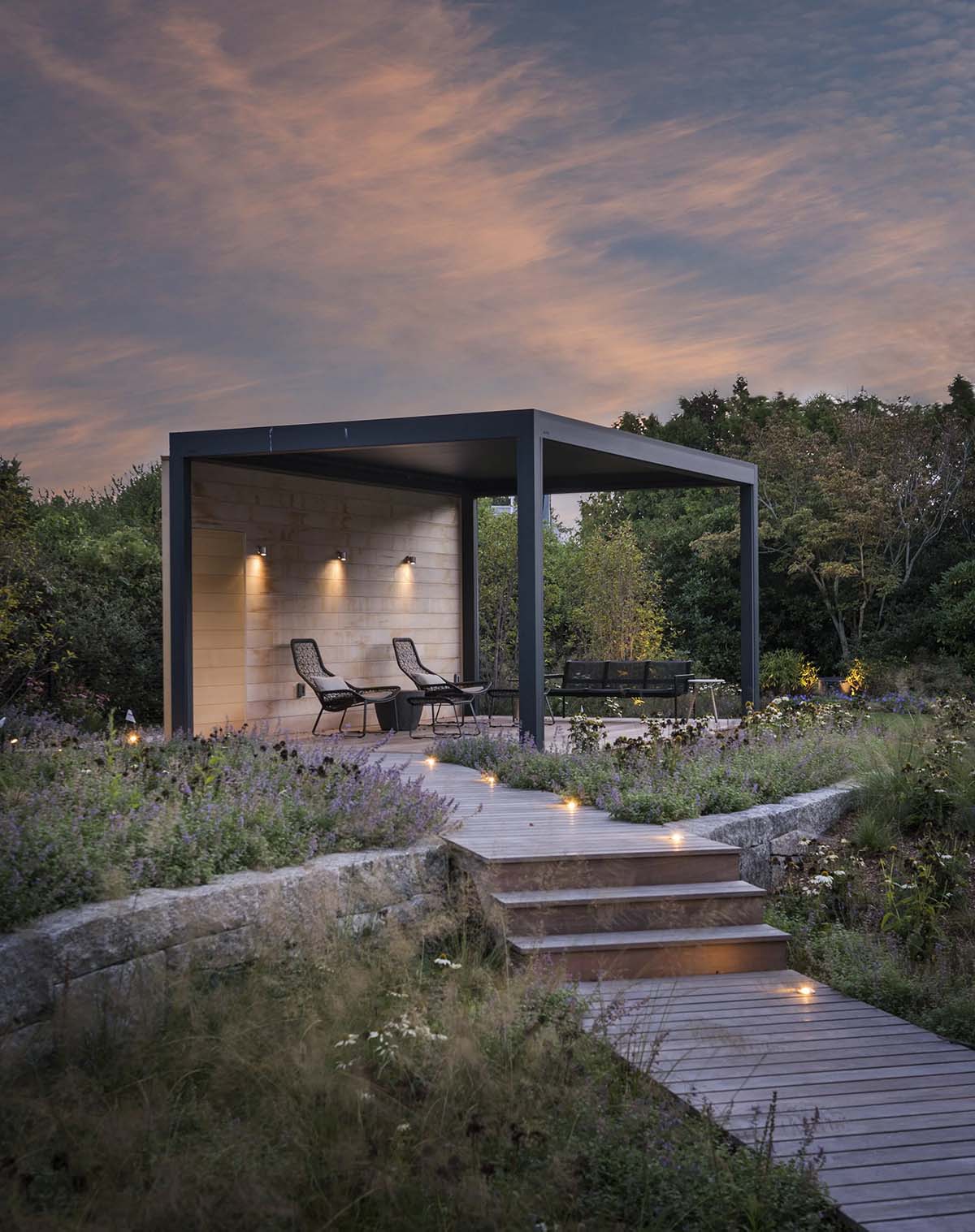 Freestanding black-framed pergola with lounge chairs, lit at dusk beside a wooden boardwalk through a wildflower meadow