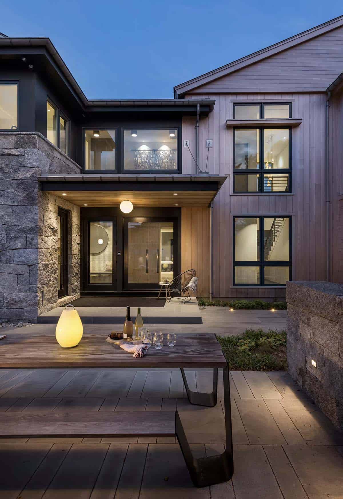 Home entrance at dusk with stone wall, glowing pendant light, and wooden dining table set with wine glasses on the patio