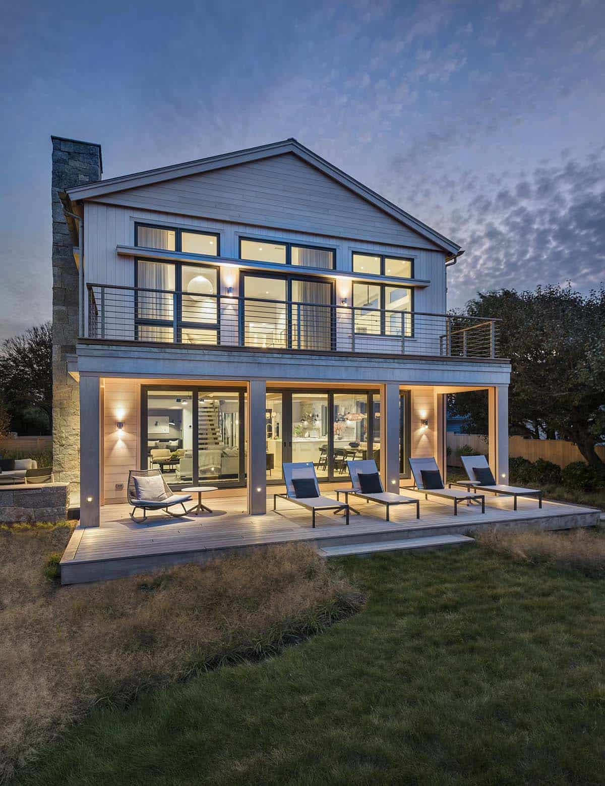 Rear view of shingled beach house at twilight, glass doors open to reveal interior lighting, with lounge chairs on the deck