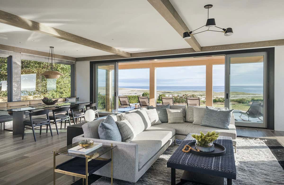 Open-concept living room with gray sectional sofa, wood beam ceiling, and folding glass doors opening to an ocean view