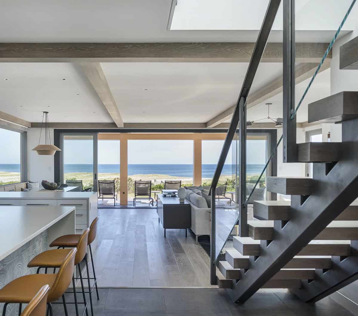 Kitchen island and living area with floating staircase, wood beams, and glass doors framing an ocean view