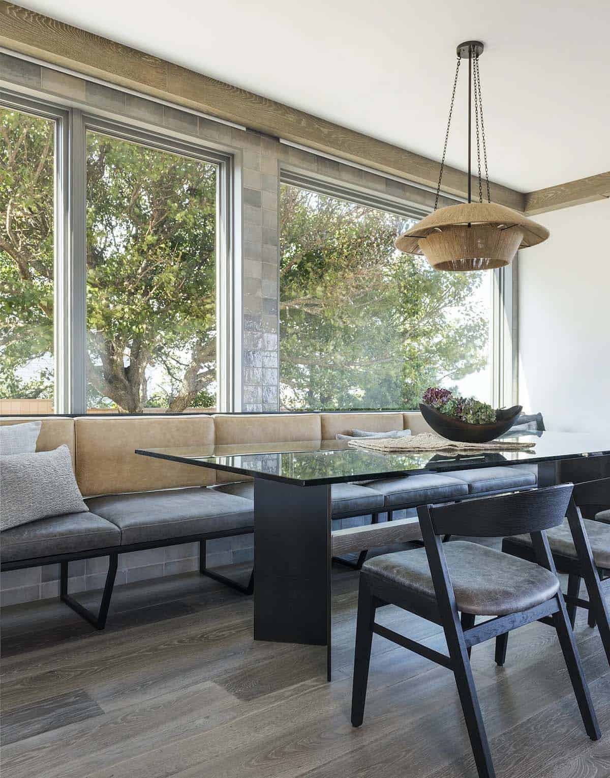 Dining banquette with tan leather cushions, glass-top table, and a woven pendant light beneath large windows