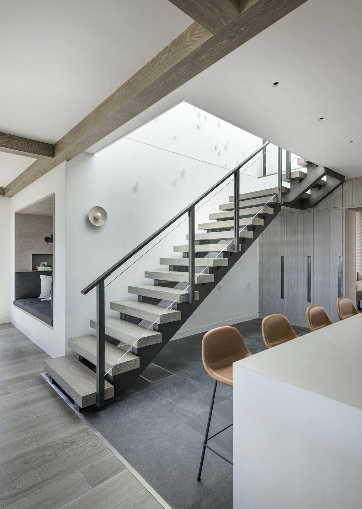Floating wood-and-steel staircase beside a kitchen island with tan leather stools and dark tile flooring