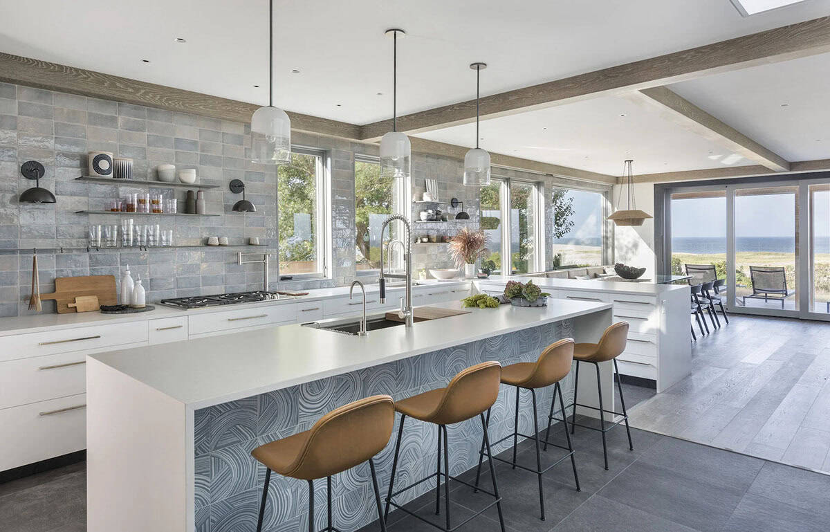 Bright kitchen with island, tan leather stools, patterned tile backsplash, and pendant lights, opening to dining area with ocean view