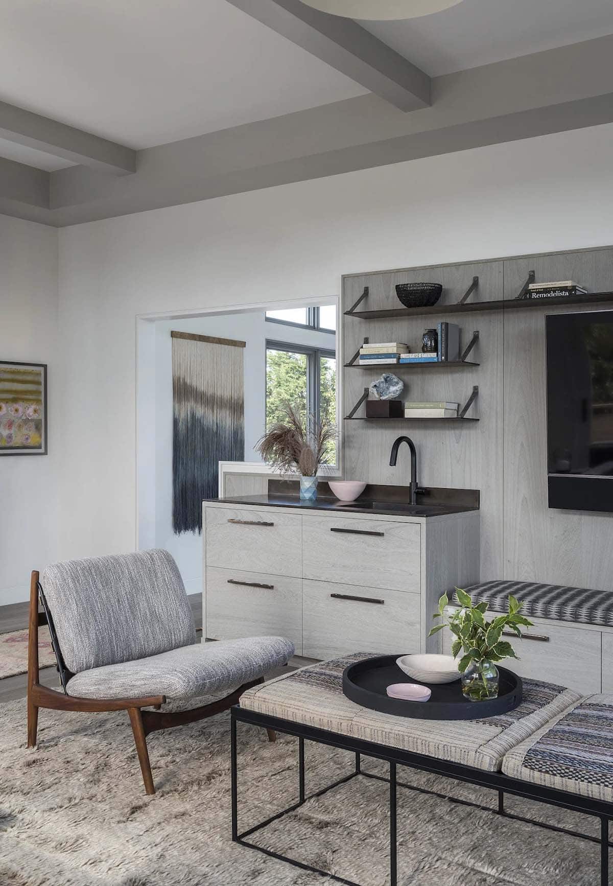 Living room seating area with mid-century chair, striped ottoman, and built-in wet bar beneath open wood shelves