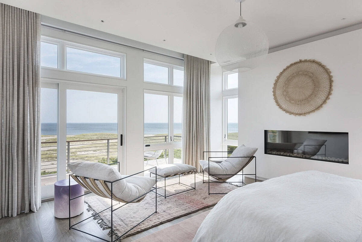 Primary bedroom with white bed, wire lounge chairs, built-in linear fireplace, and floor-to-ceiling windows facing the ocean