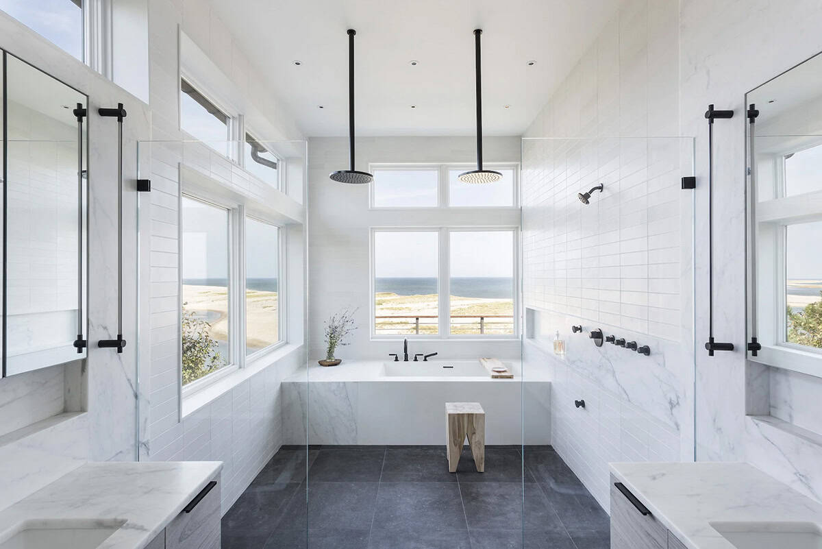 Freestanding soaking tub in a marble-tiled bathroom with dual rain shower heads and ocean-view windows