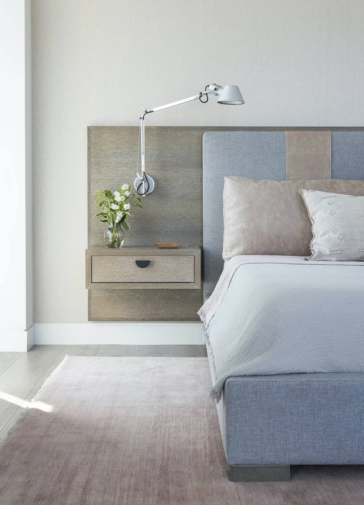 Close-up of a floating wood nightstand with an adjustable wall lamp beside a gray upholstered headboard