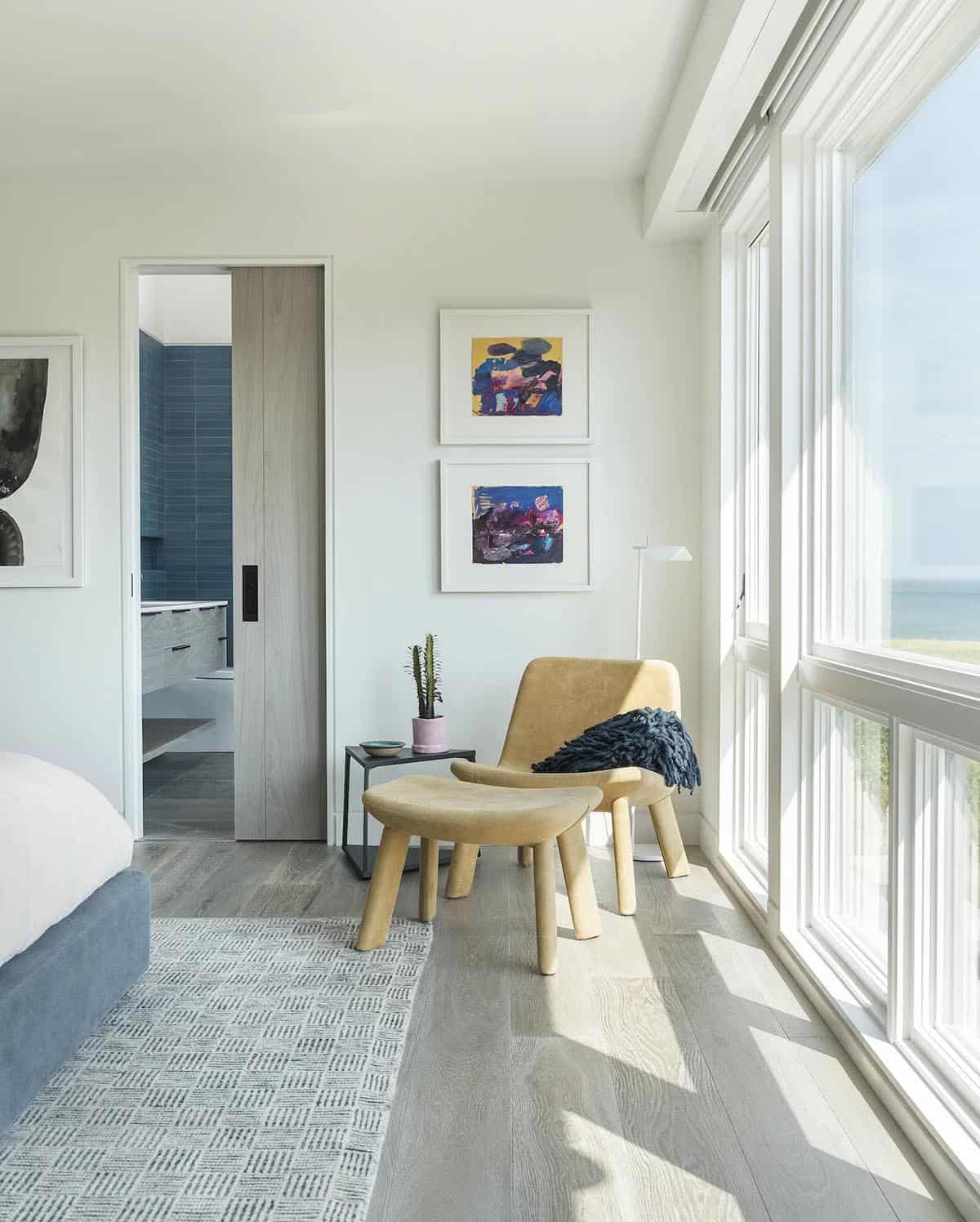 Bedroom seating nook with tan leather chair and ottoman beside floor-to-ceiling windows and framed abstract art