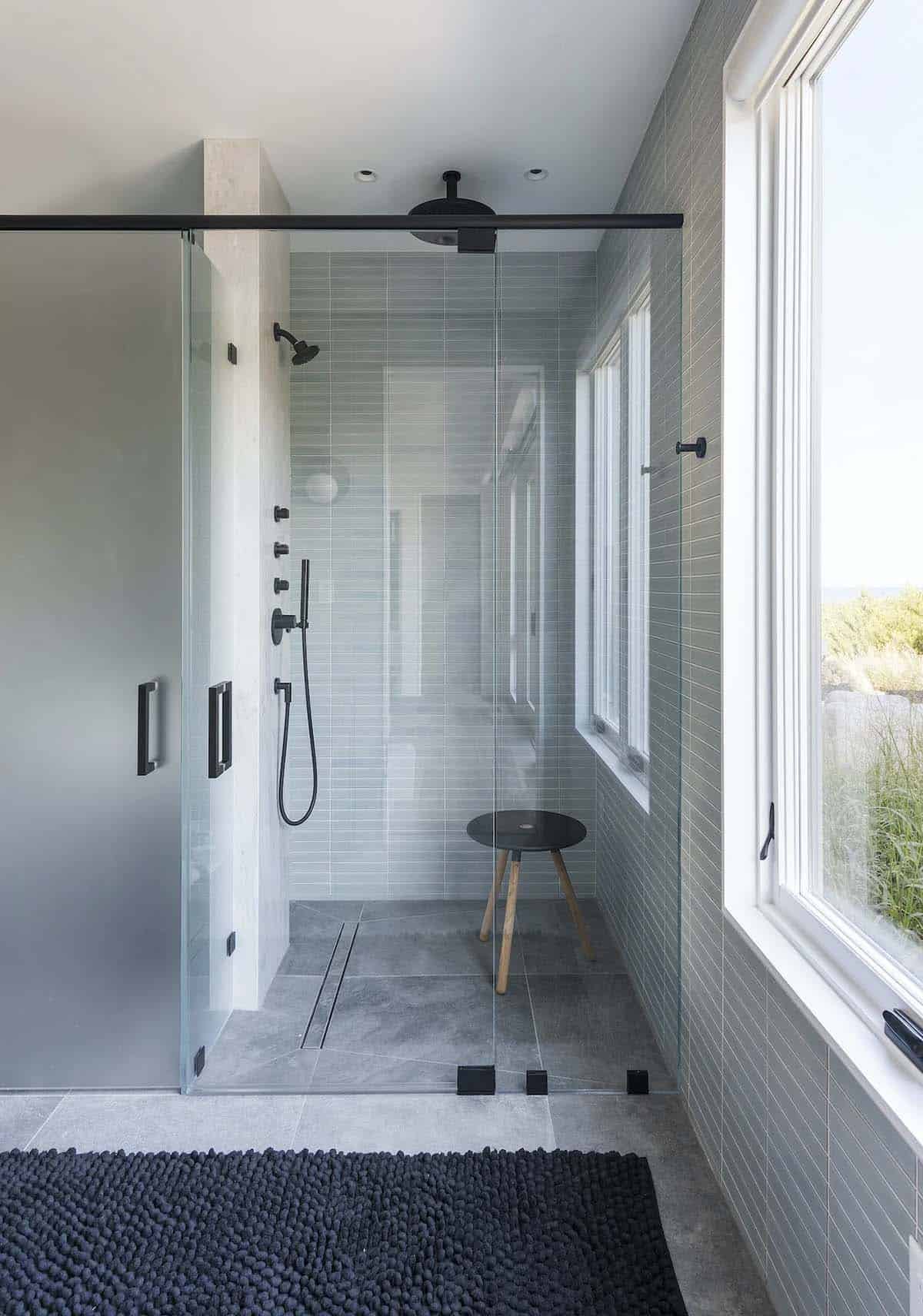 Glass-enclosed shower with black fixtures, gray linear tile, wood stool, and windows overlooking dune grass