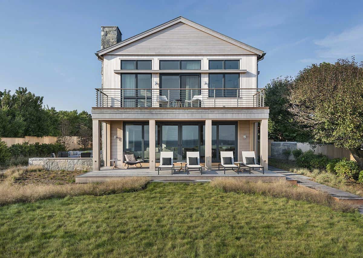 Front-facing view of shingled beach house with covered lower porch, upper balcony, and stone chimney, seen across the lawn