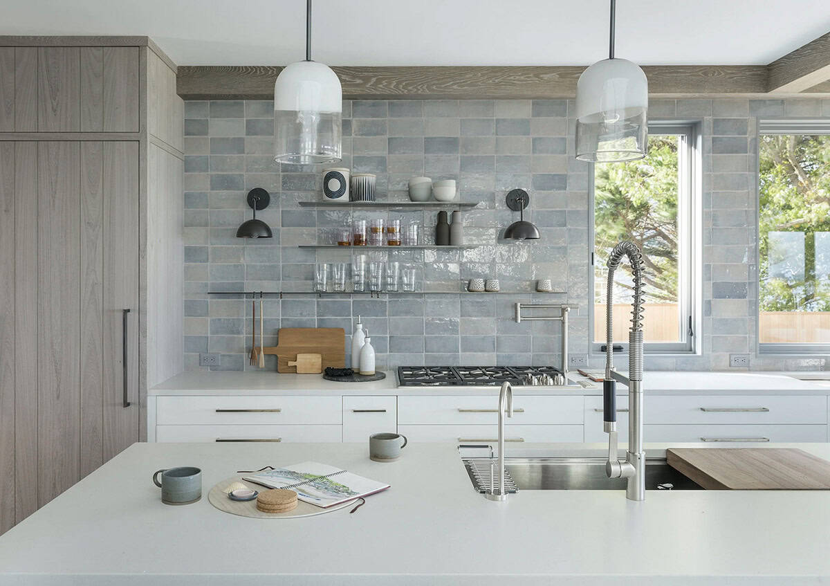 Kitchen island with pendant lights, pot filler faucet, and blue-gray zellige tile backsplash framed by windows