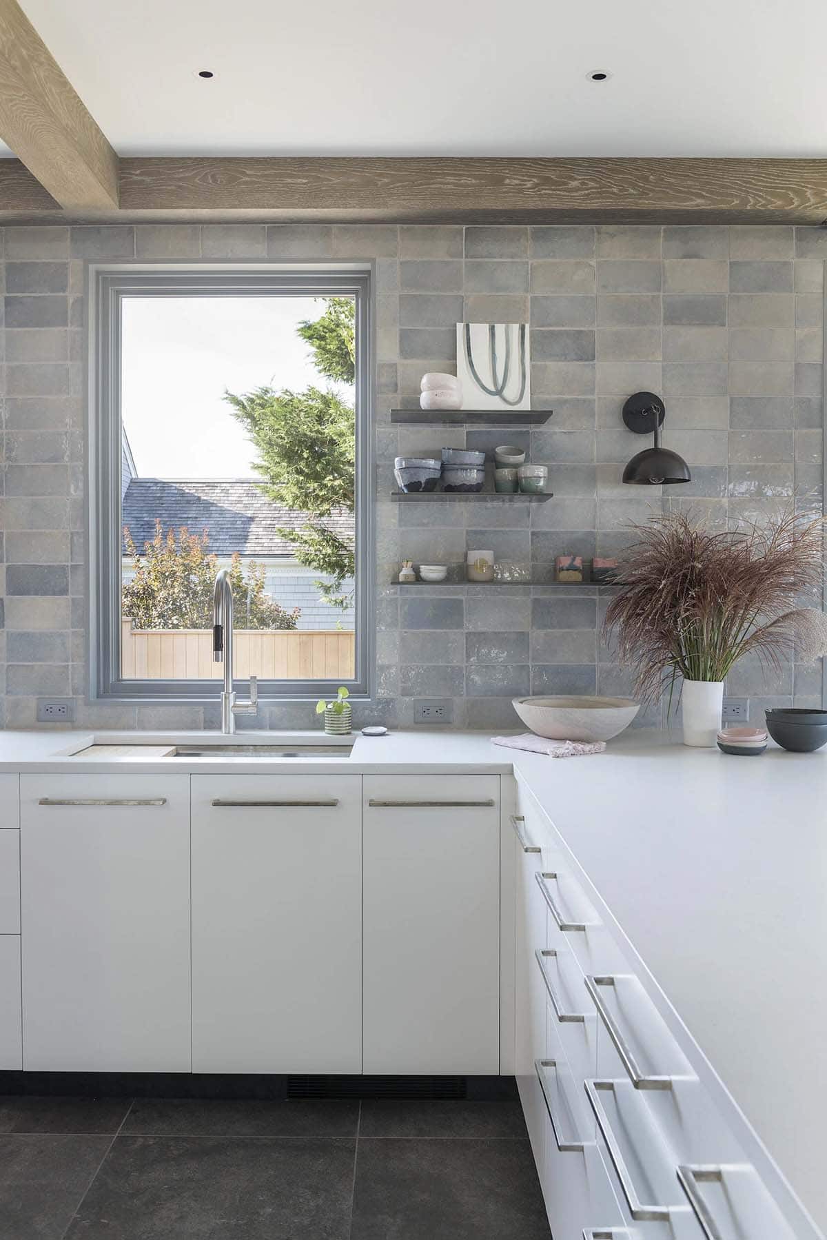 Kitchen counter corner with window over the sink, blue-gray zellige tile backsplash, open shelving, and a sconce light