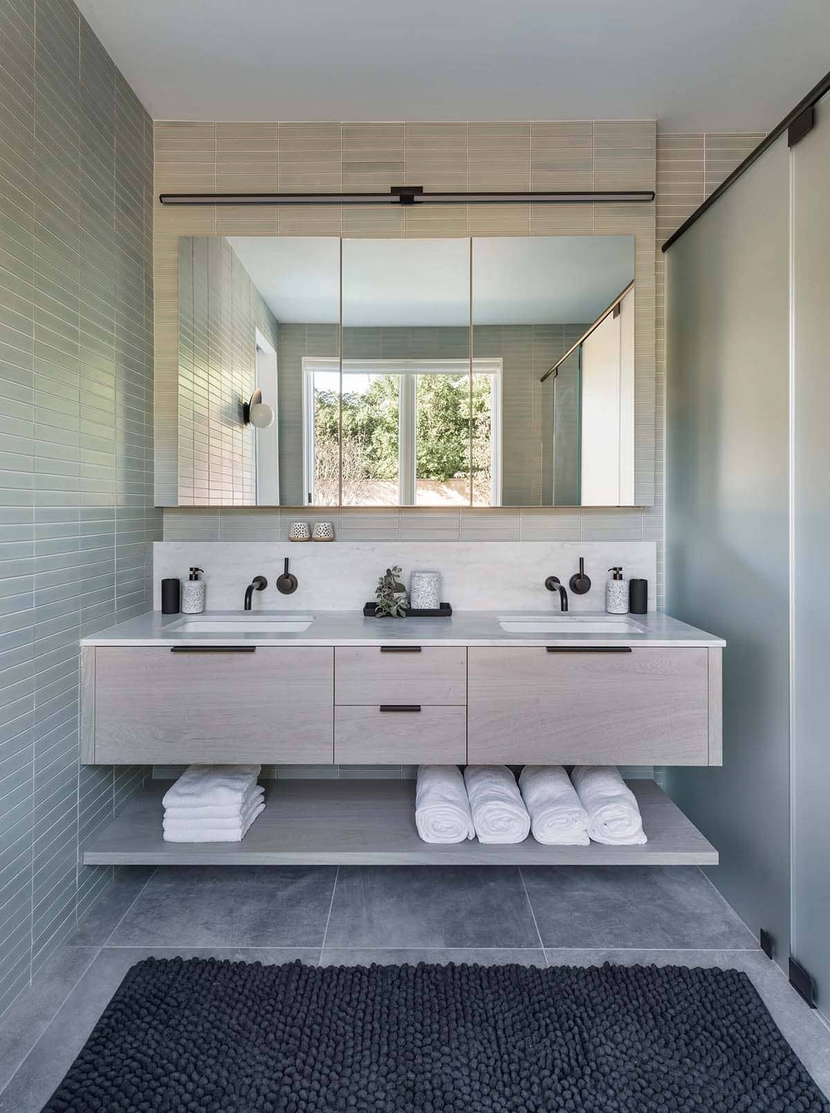 Floating double vanity with green tile walls, black fixtures, and a folded-towel display beneath a large mirror
