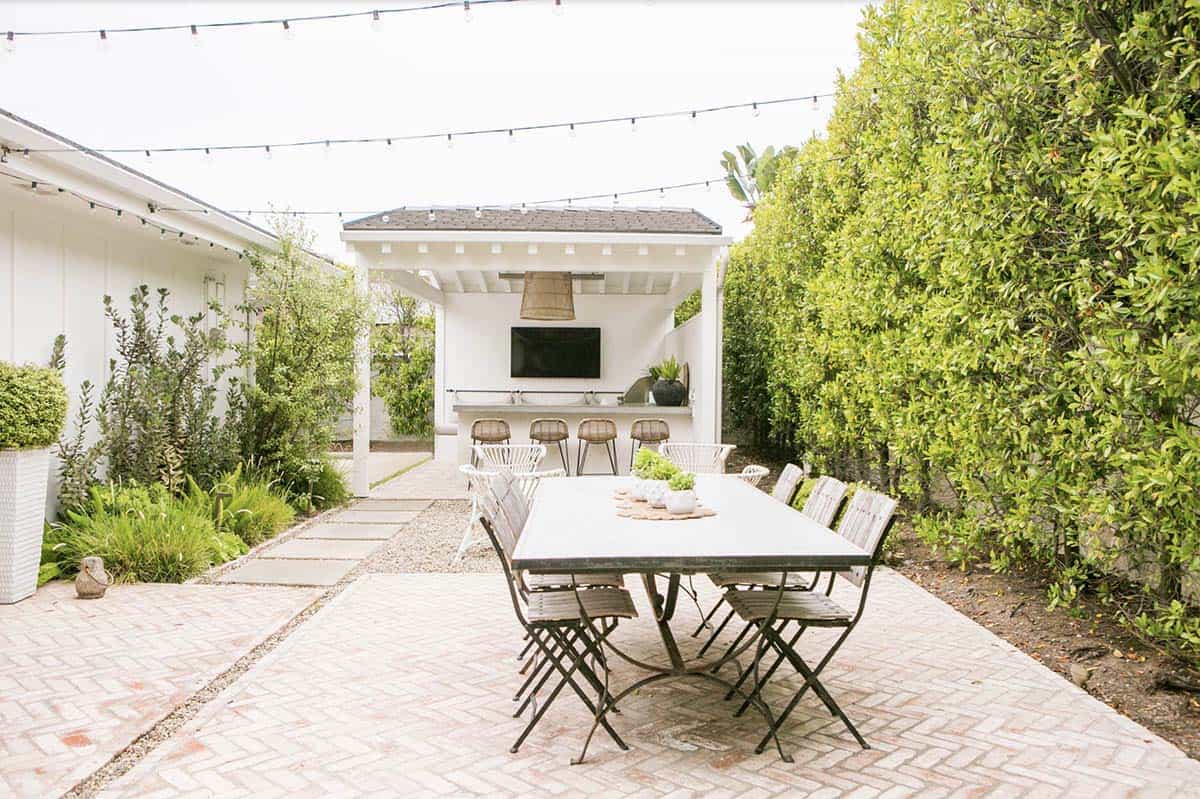 Outdoor dining table with folding metal chairs on a herringbone brick patio, string lights overhead, leading to a covered outdoor kitchen with bar seating
