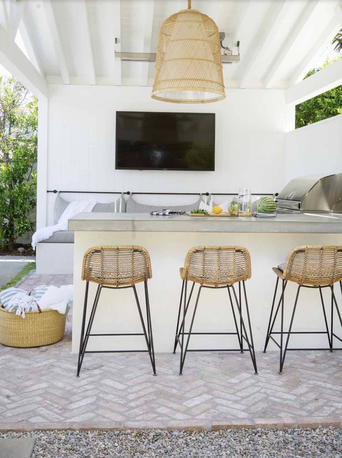 Covered outdoor kitchen with a concrete bar counter, three woven rattan bar stools, a mounted TV, and an oversized woven pendant light overhead