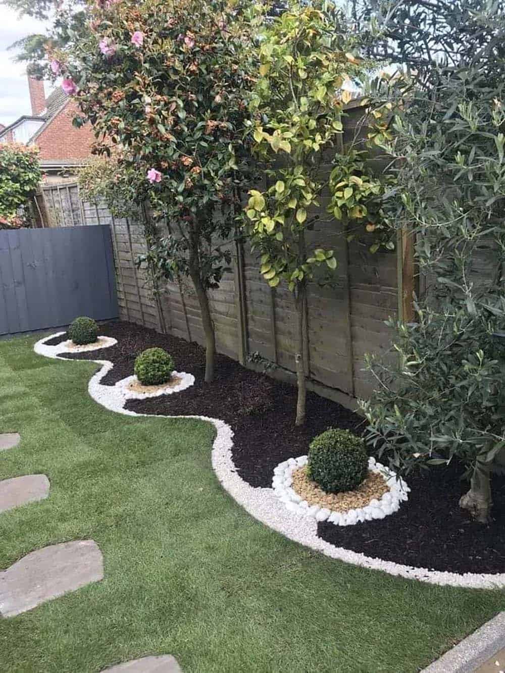Wavy white pebble border around boxwood balls and small trees along a wood fence