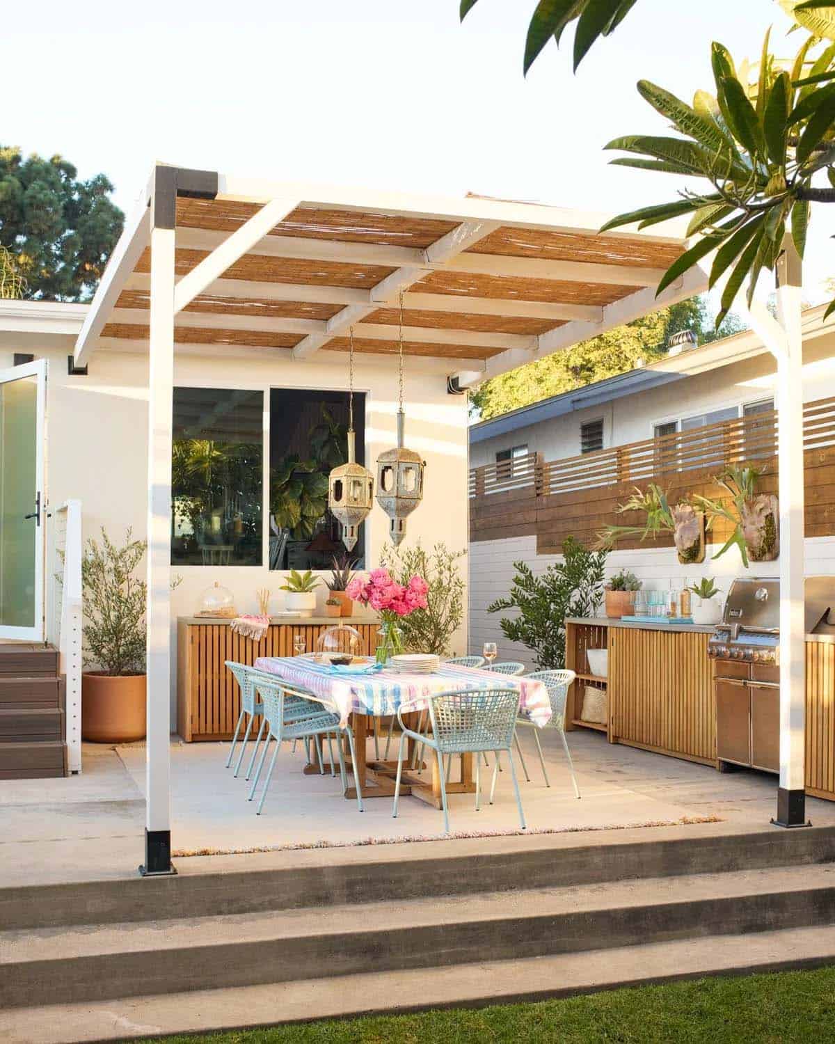 Covered patio with a white pergola over a dining table set with a striped tablecloth, pink florals, and hanging Moroccan lanterns, next to a wood-paneled outdoor kitchen with a built-in grill