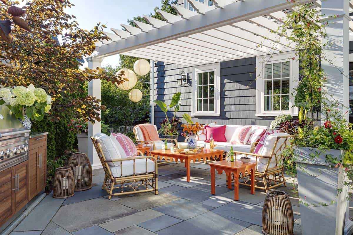 Pergola-covered patio lounge with a white sofa and bamboo chairs styled in coral and white cushions, orange lacquered coffee tables, hanging paper lanterns, and a built-in outdoor kitchen to the side