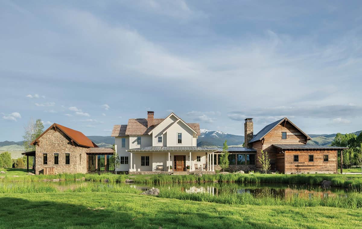 Ranch house compound with white farmhouse, stone barn, and log wing beside a pond, Montana mountains in background