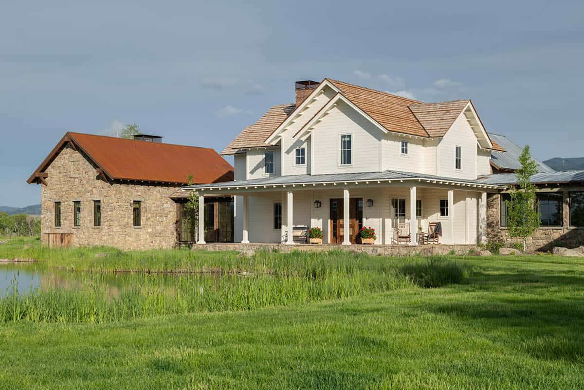 White farmhouse with wraparound porch flanked by a stone building with rust-colored roof, pond in foreground