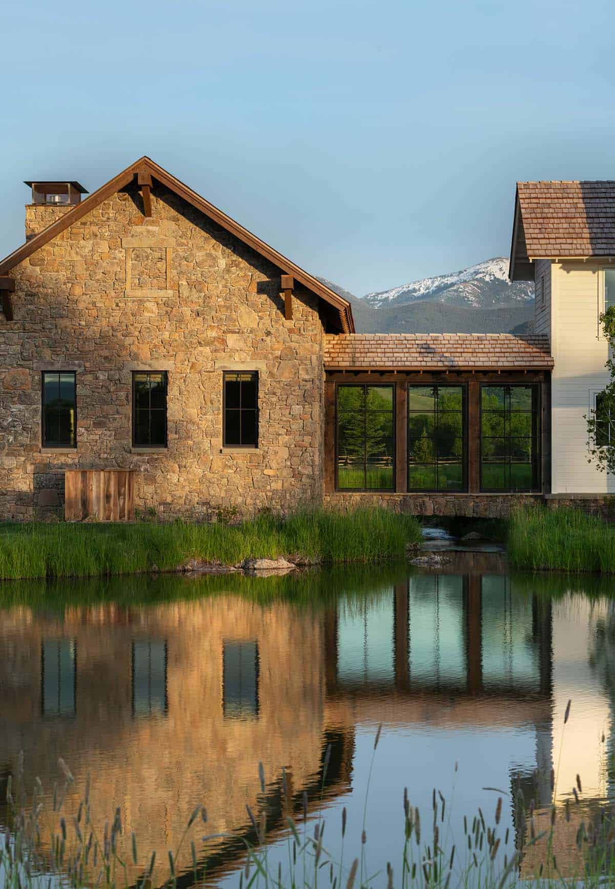 Stone gable-end building with glass sunroom addition reflected in a still pond at golden hour, mountains beyond