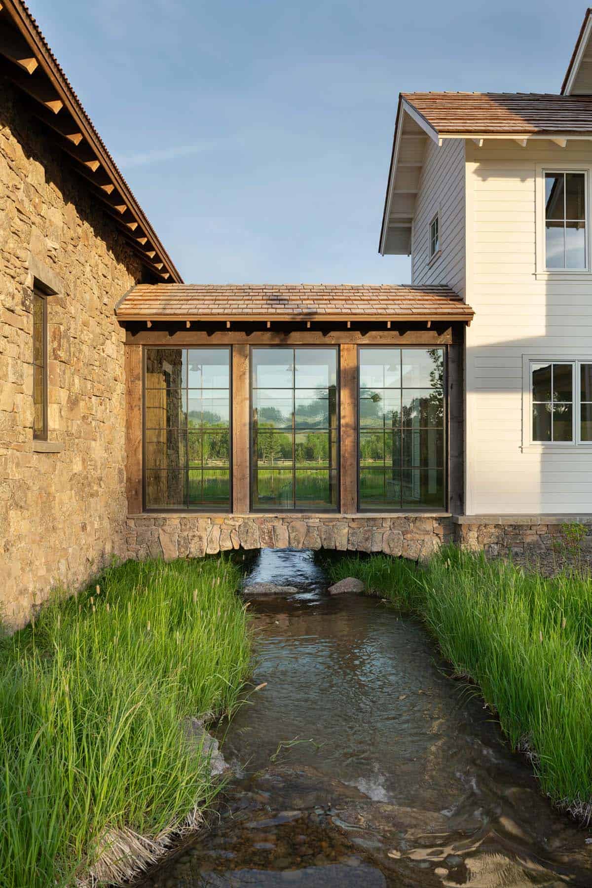 Glass-walled breezeway connecting stone building to white farmhouse, built over a small stone creek crossing