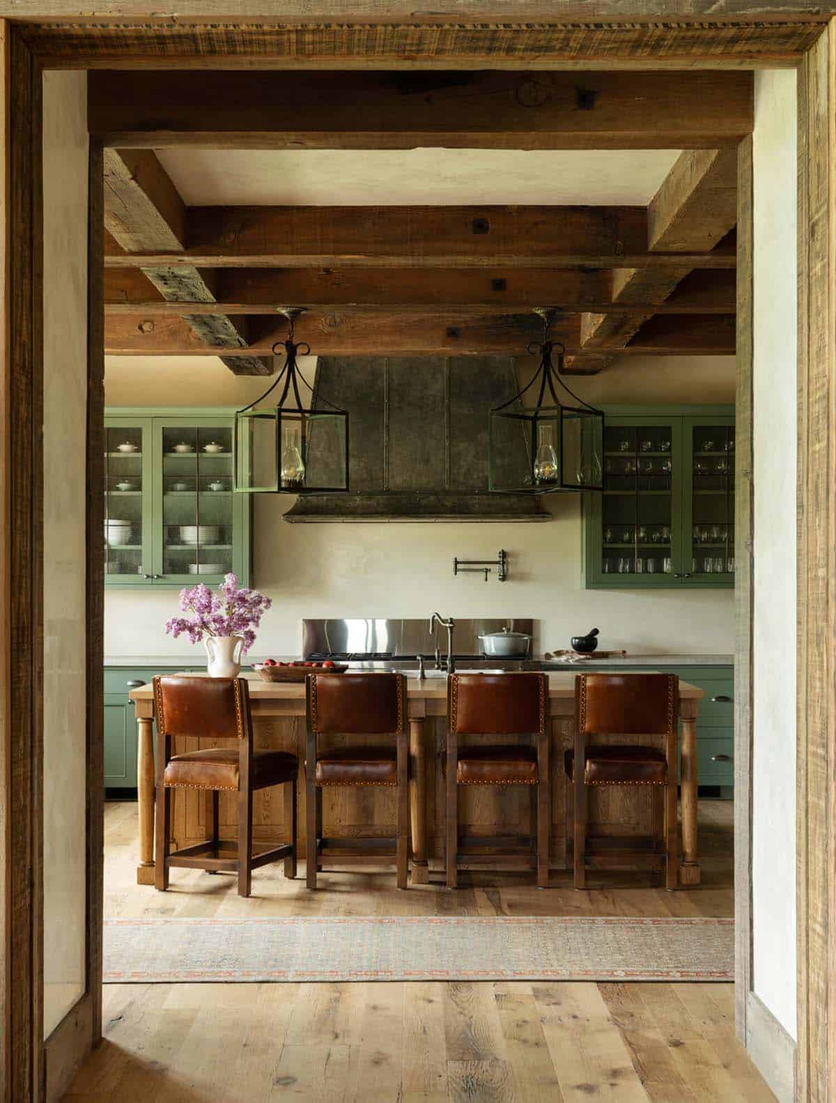 View through reclaimed wood beam doorway into kitchen with sage-green cabinets and leather counter stools