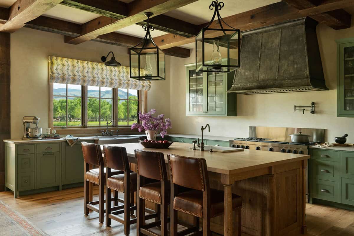 Farmhouse kitchen with sage-green cabinetry, wood island, leather stools, and dark metal range hood under exposed beams